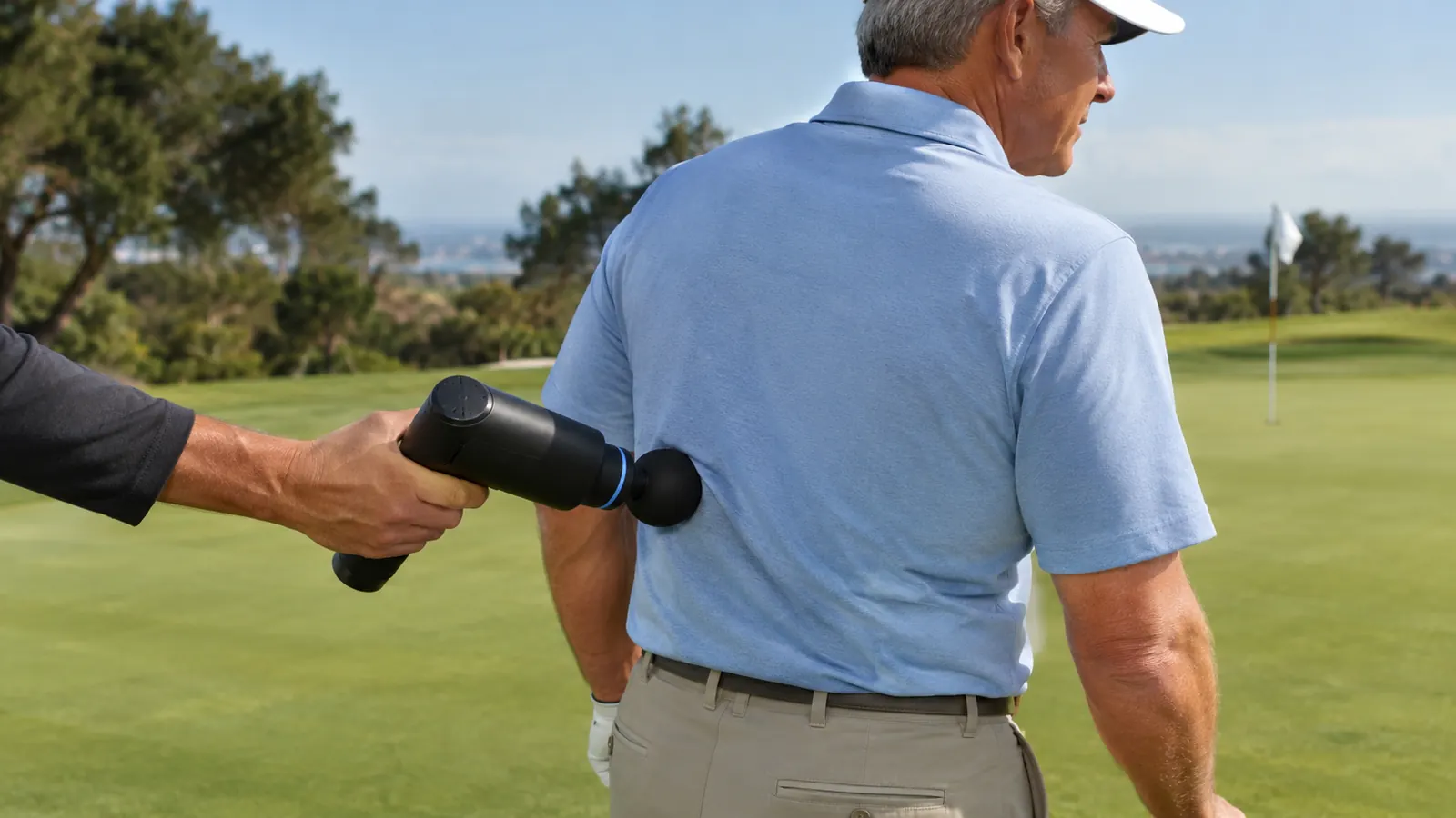 A hand holding a massage gun against the lower back of a golfer standing on a practice green
